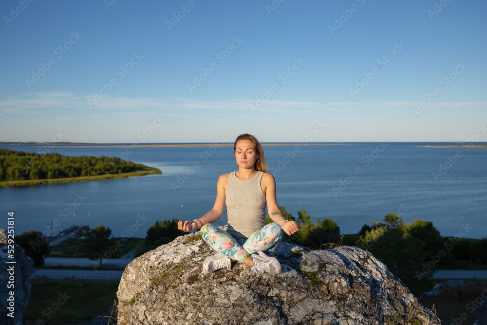 Blondie woman in lotus yoga pose are sitting on the rock near sea cost