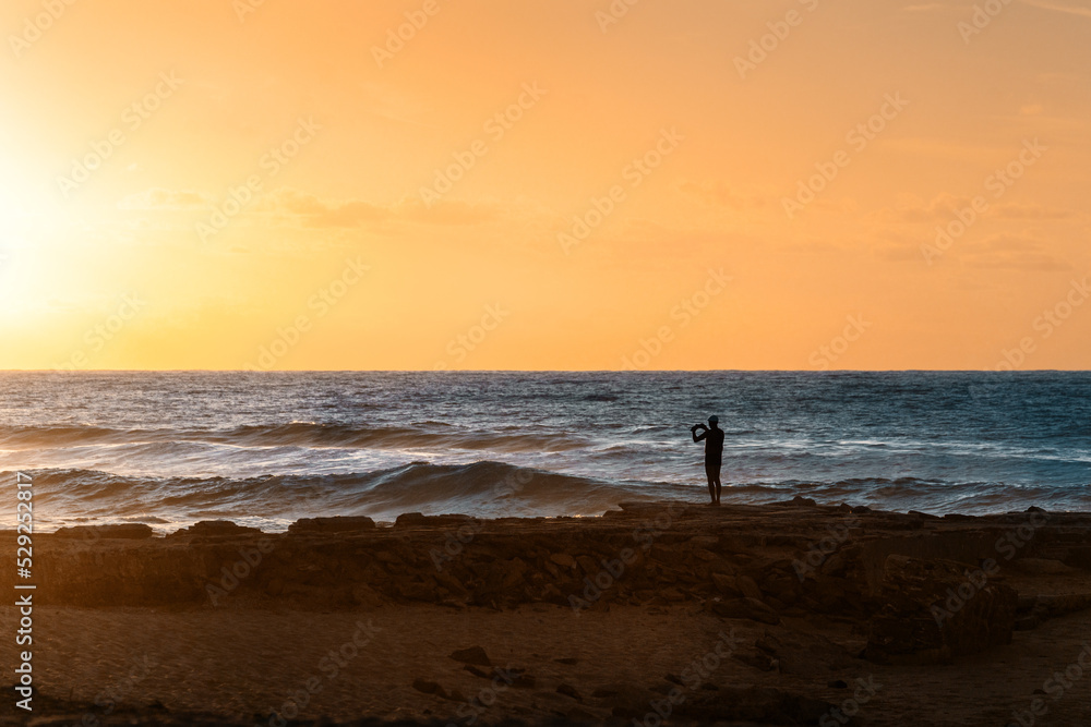 Mann steht am Strand Spiaggia di Scivu auf Sardinien Italien und fotografiert den Sonnenuntergang