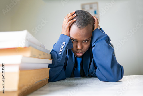 Giovane ragazzo nero in giacca e cravatta depresso seduta con i gomiti appogiate sulla scrivania e le mani intorno alla testa e guarda i libri  photo