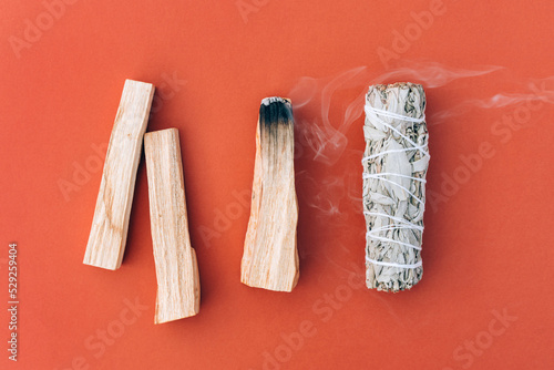 Smoldering incense. White sage and palo santo sticks in a row over orange red background. Bundle for meditation and room fumigation. Selective focus. Flat lay style