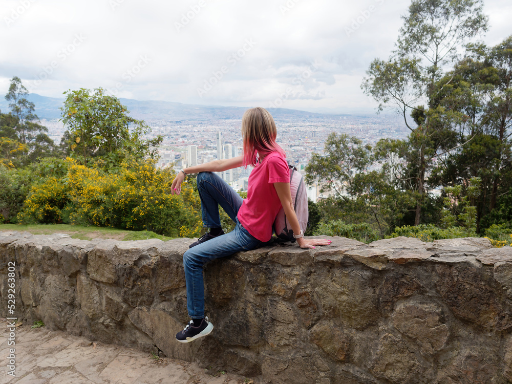 Woman sitting on stone fence, on top of mountain looking on city.