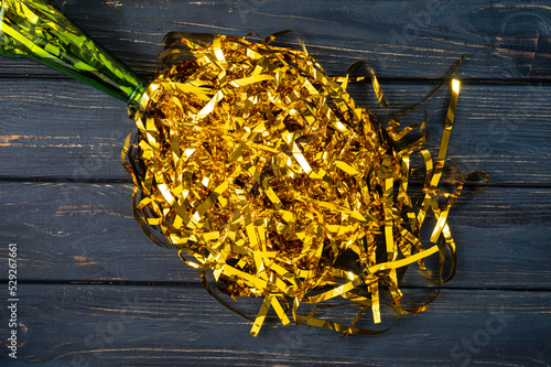 Top view of the neck of a green champagne bottle with a splash of golden tinsel splash. Golden shiny serpentine ribbons, sequins on gray wooden table. Glass bottle with glittering golden tinsel.