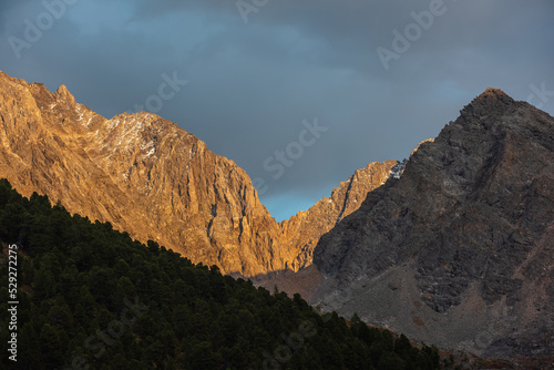 Awesome view to large mountain range with sharp rocks in golden sunlight under cloudy sky. Sunlit gold high mountain ridge with under dramatic gray sky in changeable weather. Rainy clouds in mountains