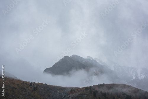 Dark atmospheric landscape with high mountain silhouettes in dense fog in rainy weather. Snowy rocky mountain top above hills in thick fog in dramatic overcast. Black rocks in low clouds during rain. © Daniil