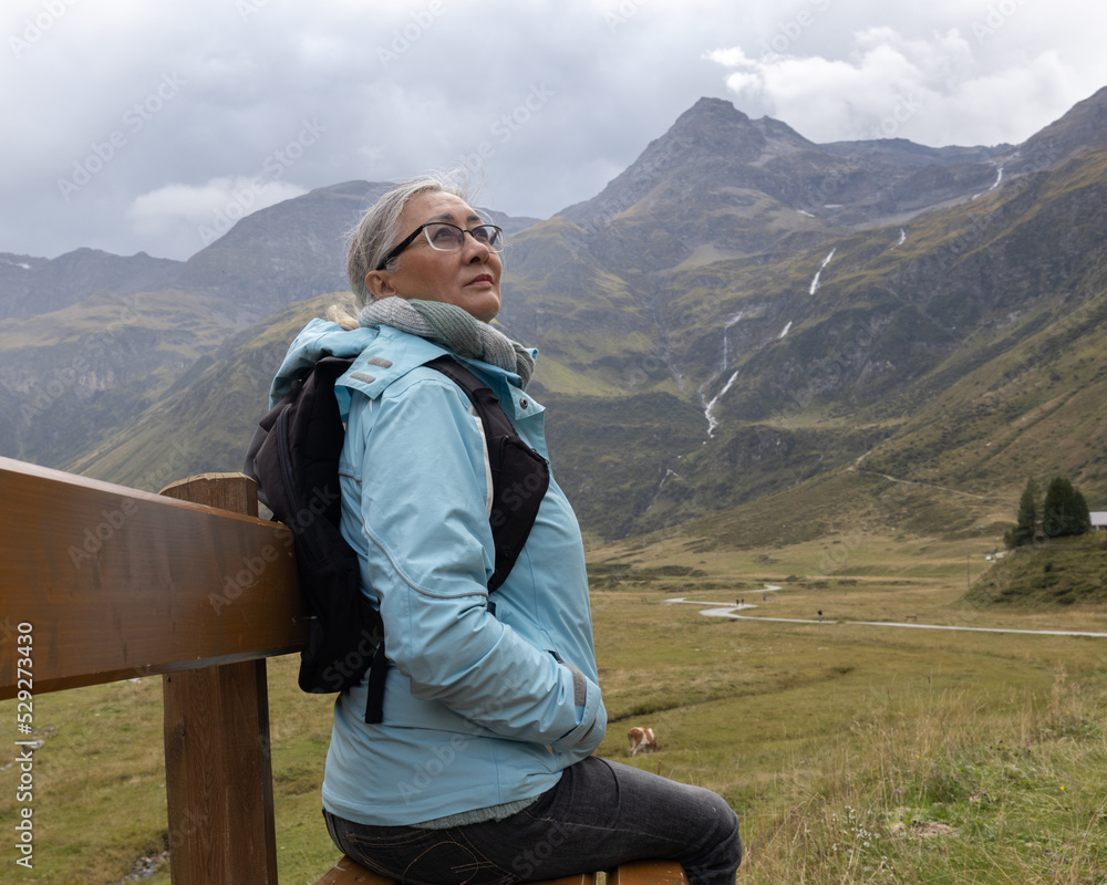 A gray-haired woman in a windbreaker with a backpack sits on a wooden bench in an alpine valley.