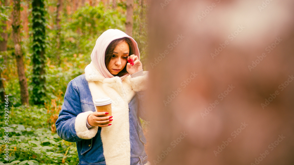 Fototapeta premium Young woman rubs her eye, holding paper cup with coffee, standing in forest.