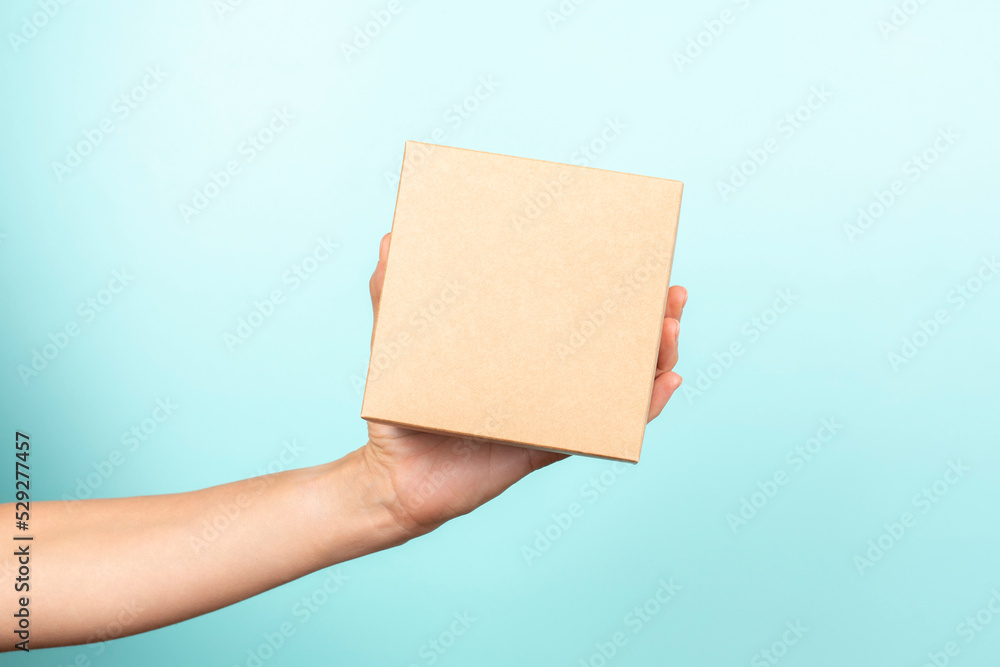 Woman hand show small brown rectangular cardboard box on light blue background. Mockup parcel box. Packaging, shopping, delivery, present, gift concept Woman hand show small brown rectangular cardboard box on light blue background. Mockup parcel box. Packaging, shopping, delivery, present, gift concept