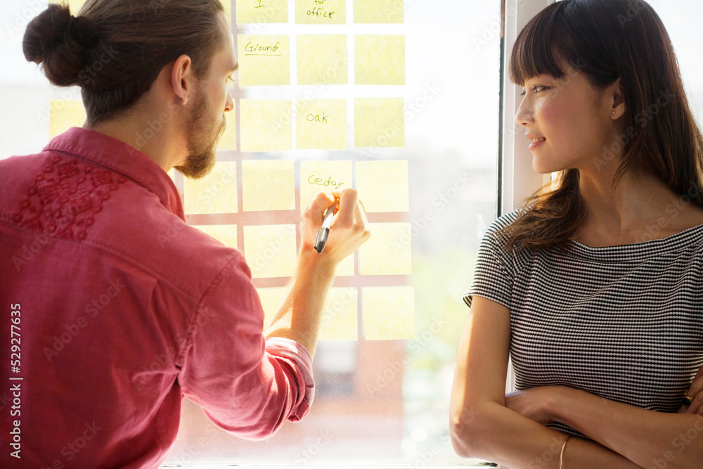 Businesswoman looking at colleague writing on adhesive note in creative office Businesswoman looking at colleague writing on adhesive note in creative office