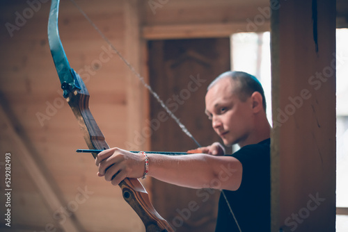 A man in a shooting gallery holds a bow in his hands and aims. The bowstring is being pulled. Front view.