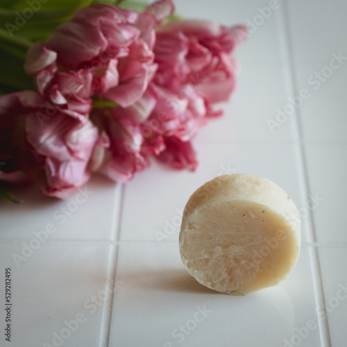 Textured round bar of hard soap with a rough texture close-up against the background of white tiles and a bouquet of pink tulips.