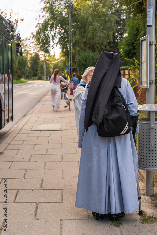 A Catholic nun in monastic garb at a bus stop talking to an elderly woman in Krakow, Poland, with bus passengers walking down the sidewalk ahead