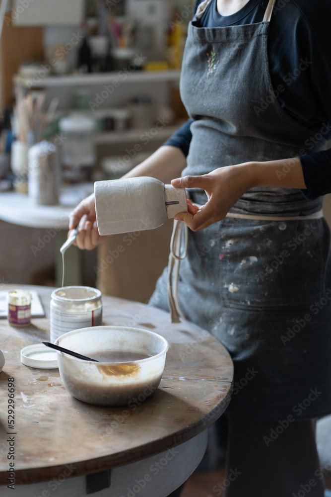 Young craftswoman in black apron paints clay vase with paintbrush after baking and handmade shaping in studio. Woman enjoys painting earthenware vase in workroom for handmade pottery shop closeup