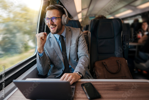 Handsome businessman is having a good time while traveling by high-speed train. He is using laptop computer and wireless headphones for online communication, gaming and entertainment. photo