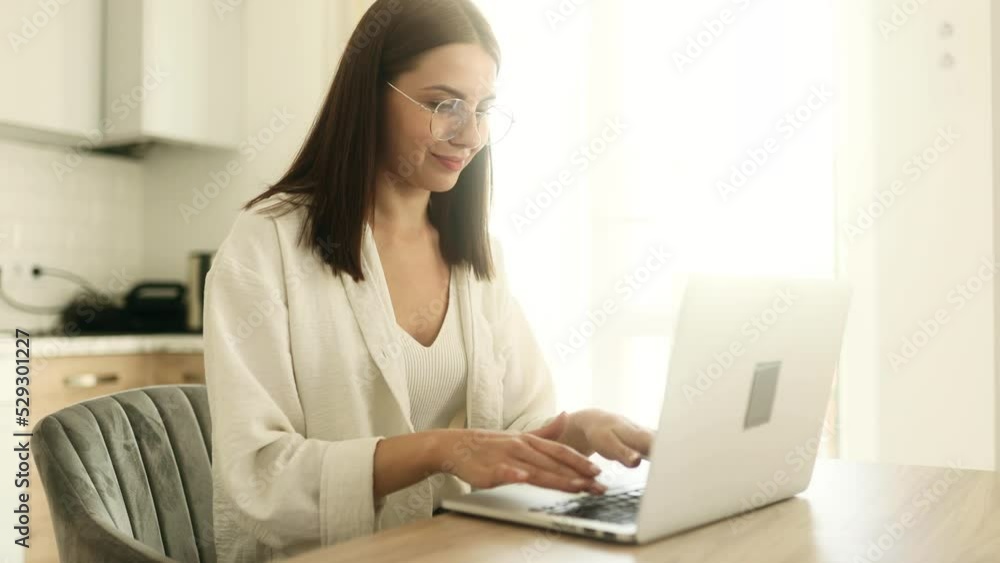 Smiling young woman in eyeglasses using laptop in the kitchen. Young brunette with computer, freelancer working at home. Smiling young woman in eyeglasses using laptop in the kitchen. Young brunette with computer, freelancer working at home.