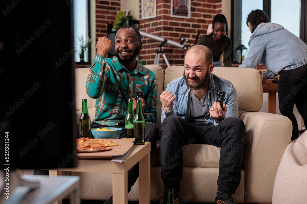 Fototapeta premium Cheerful people celebrating video games win at house gathering, using tv console to play competition. Men feeling happy about winning, having fun wiht friends at party with beer.