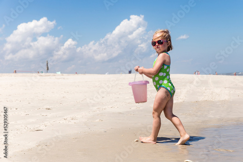 Full length of girl wearing one piece swimsuit and sunglasses while carrying bucket at beach during sunny day photo