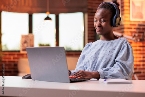 Smiling student in headphones attending online classes on laptop at home. African american woman watching educational video in modern room with big windows and beautiful warm sunset light
