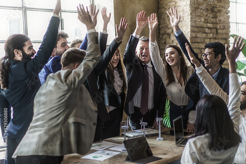 Successful multiethnic group of business people cheering raising hands standing around the meeting table - multiracial businessmen and businesswoman having fun - business lifestyle concept
