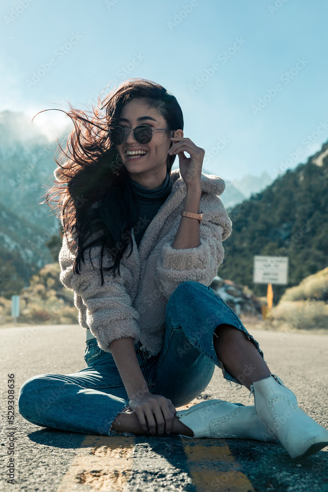 Full length of cheerful young woman wearing sunglasses while sitting on road during sunny day Full length of cheerful young woman wearing sunglasses while sitting on road during sunny day