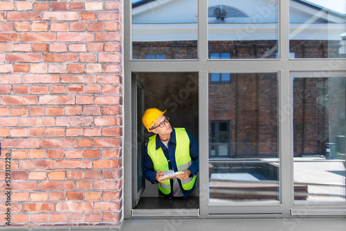 construction business and building concept - male builder in helmet and safety west with clipboard and pencil looking out window