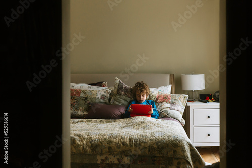 Boy using tablet computer while reclining on pillows in bedroom at home