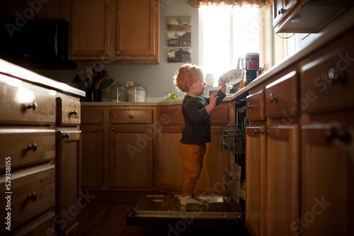 Young boy helps do chores standing on dishwasher putting dishes away photo