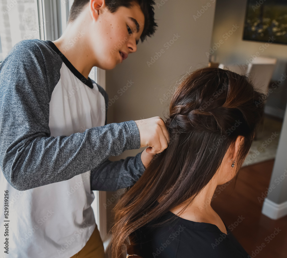 Teenage boy braiding his mother's hair near a window.