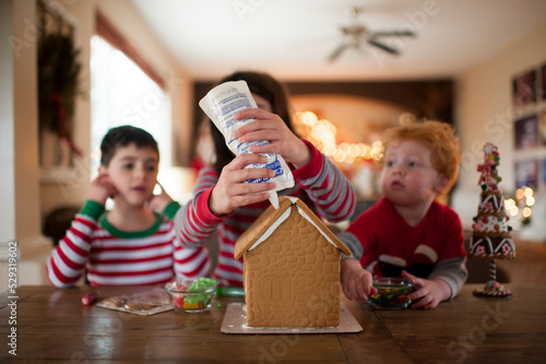 Brothers watch as sister adds frosting to a holiday gingerbread house