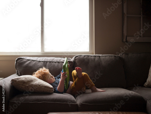 Young boy lays on couch at home watching his tablet by window photo