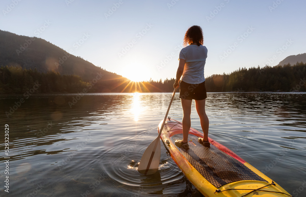 Adventurous Woman Paddling on a Paddle Board in a peaceful lake. Sunny Sunset. Hicks Lake, Sasquatch Provincial Park near Harrison Hot Springs, British Columbia, Canada. Adventurous Woman Paddling on a Paddle Board in a peaceful lake. Sunny Sunset. Hicks Lake, Sasquatch Provincial Park near Harrison Hot Springs, British Columbia, Canada.