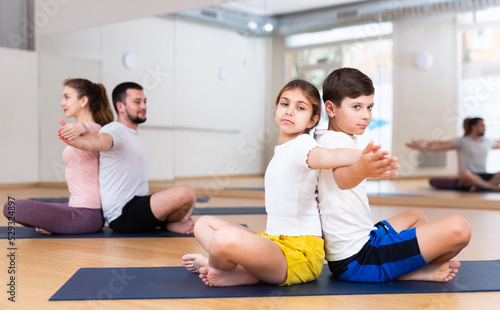 Tween girl with brother sitting on mat in modern yoga studio during family workout, doing exercises in pair ..