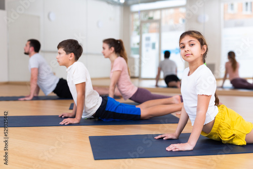 Girl looking at camera while doing cobra pose with her brother and parents on yoga family training.