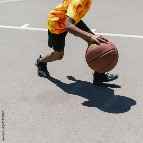Unrecognizable dark skinned kid with a yellow sports uniform plays basketball on a sidewalk on a urban street. There are no trademarks in the shot.