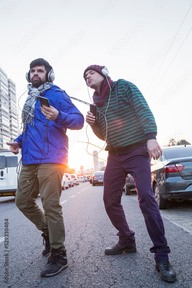 Two gay guys listening music from smartphone with headphones while dancing near the traffic on the street . Golden Hour. LGBTQ people life. Copy space. Vertical.