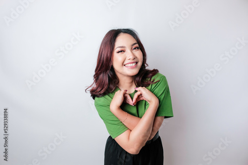 An attractive young Asian woman wearing a green t-shirt feels happy and a romantic shapes heart gesture expresses tender feelings