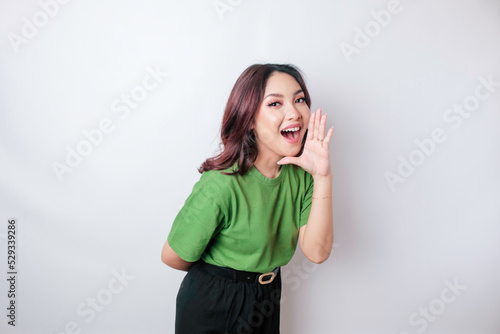 Young beautiful woman wearing a green t-shirt shouting and screaming loud with a hand on her mouth. communication concept. © Reezky