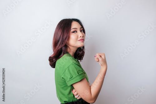 Excited Asian woman wearing a green t-shirt showing strong gesture by lifting her arms and muscles smiling proudly