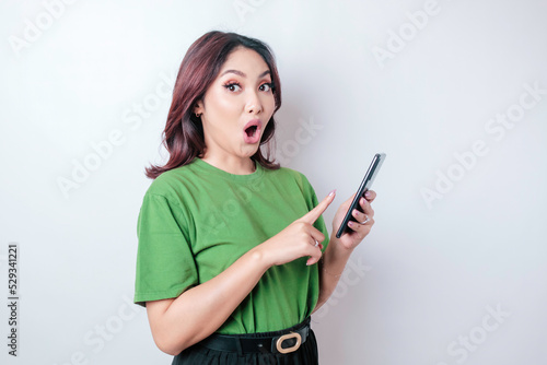 Surprised Asian woman wearing green t-shirt pointing at her smartphone, isolated by white background