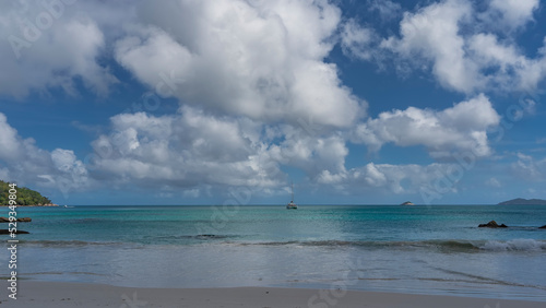 Serene seascape. The waves of the turquoise ocean roll onto the sandy beach. Boulders in the water. Yachts in the distance. Picturesque cumulus clouds in the blue sky. Seychelles. Praslin Island.