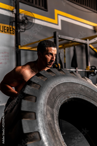 High quality photo. Muscular shirtless Hispanic man lifting a big tire in a gym training with all his might.