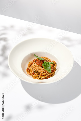Vegetarian pasta with basil and tomatoes on white plate. Tomato pasta in summer menu with hard shadows Spaghetti with tomatoes on light background. Vegan food - spaghetti marinara