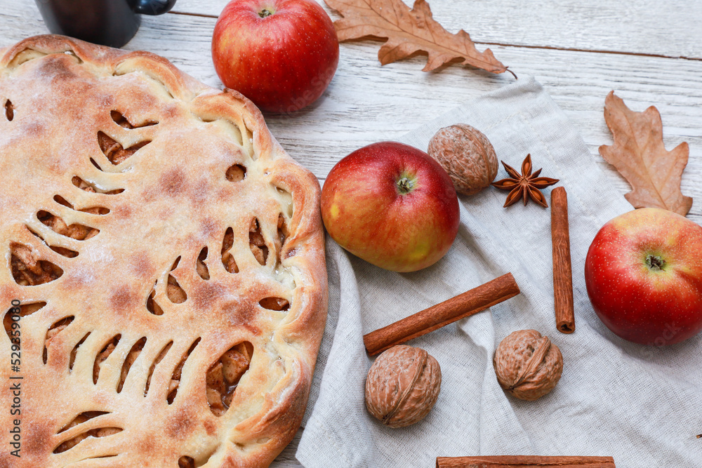 Whole apple pie on linen napkin, fresh fruits, nuts, spices, dry autumn leaves on white wooden rustic table background. Top view, close up. Homemade pastry, traditional dessert, bakery food