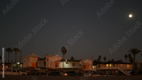 Palm trees silhouettes and full moon in twilight sky, California beach, USA. Beachfront houses or homes on coast in evening, fullmoon on pacific ocean shore in dusk. Lifeguard tower. Light in windows.
