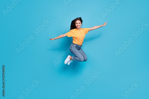Full length photo of adorable pretty girl dressed yellow t-shirt arms sides jumping high isolated blue color background