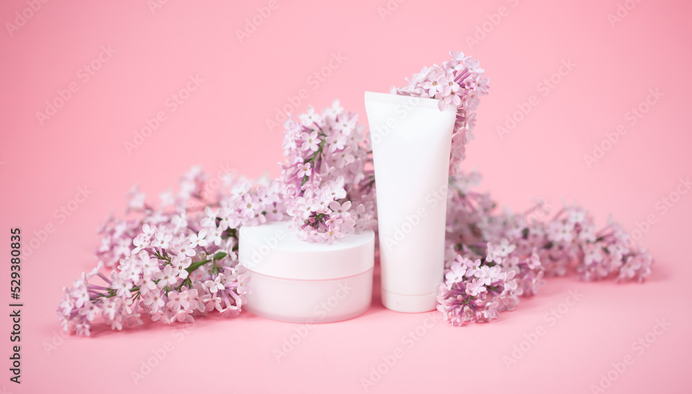 Face and body skin care. A set of cosmetic creams and balms in white tubes and cans on a coral background with sprigs of lilac flowers. Spa treatments for home care. Home rejuvenation and moisturizing Face and body skin care. A set of cosmetic creams and balms in white tubes and cans on a coral background with sprigs of lilac flowers. Spa treatments for home care. Home rejuvenation and moisturizing