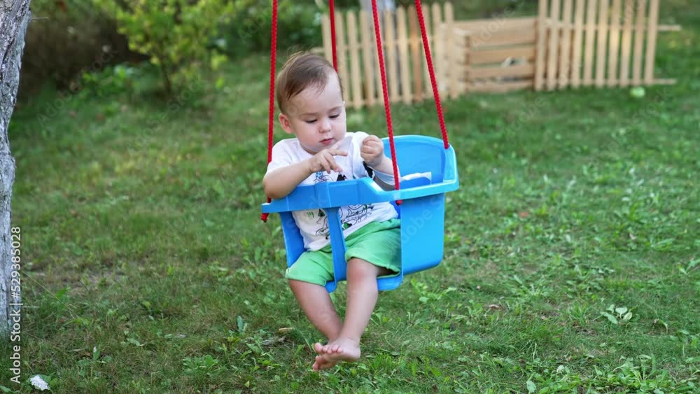 Lovely boy in white t-shirt and green shorts outdoors. Little baby swaying in the plastic swing tied to the tree. Garden backdrop. Lovely boy in white t-shirt and green shorts outdoors. Little baby swaying in the plastic swing tied to the tree. Garden backdrop.