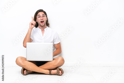Young handsome man with a laptop sitting on the floor isolated on white background thinking an idea pointing the finger up © luismolinero