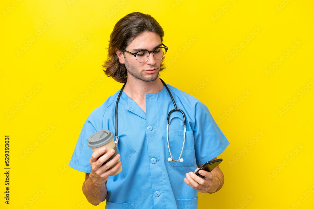 Young surgeon caucasian man isolated on yellow background holding coffee to take away and a mobile Young surgeon caucasian man isolated on yellow background holding coffee to take away and a mobile