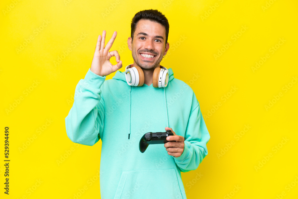 Young handsome man playing with a video game controller over isolated wall showing ok sign with fingers Young handsome man playing with a video game controller over isolated wall showing ok sign with fingers