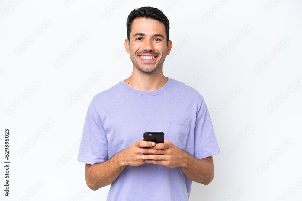 Young handsome man over isolated white background looking at the camera and smiling while using the mobile